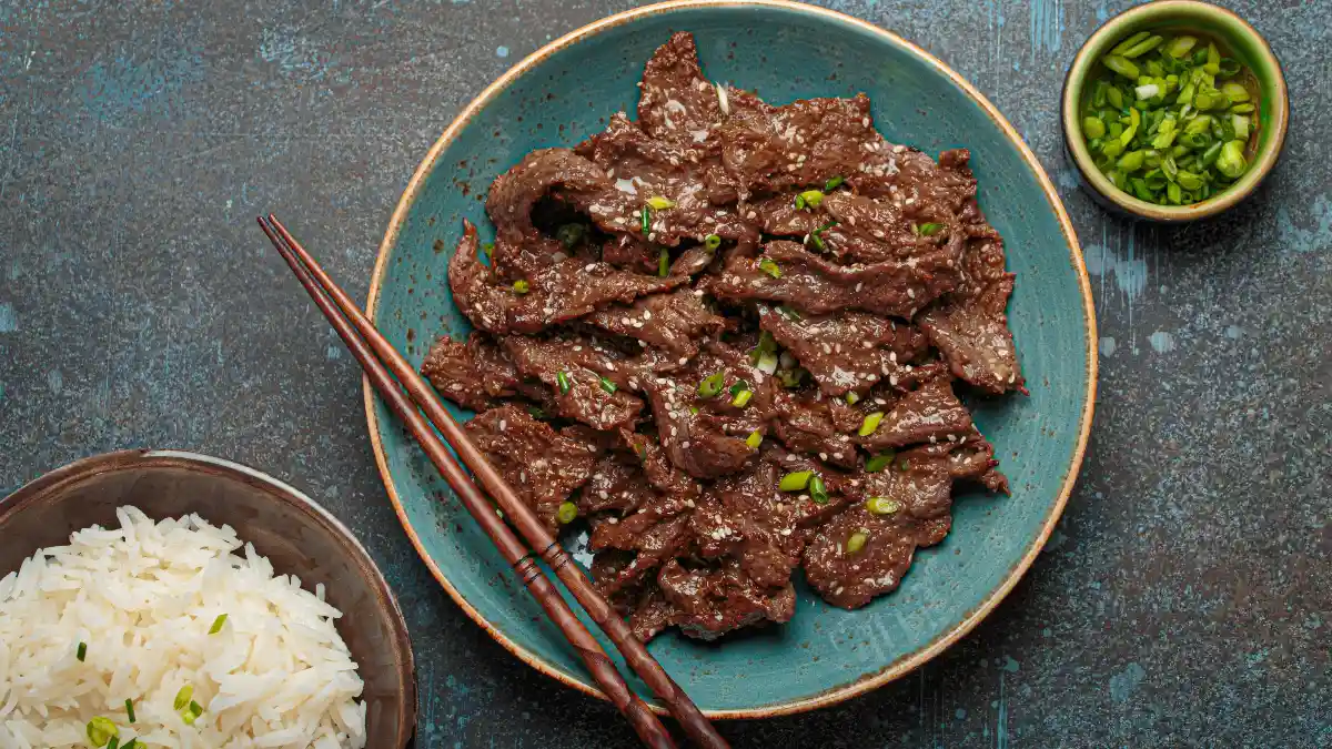 Tiras de carne servida em uma tigela azul, com um par de hashis e com outros acompanhamentos.