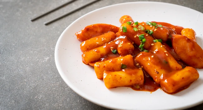 Tteokbokki servido em um prato branco, redondo e raso, ao fundo, um par de hashis.